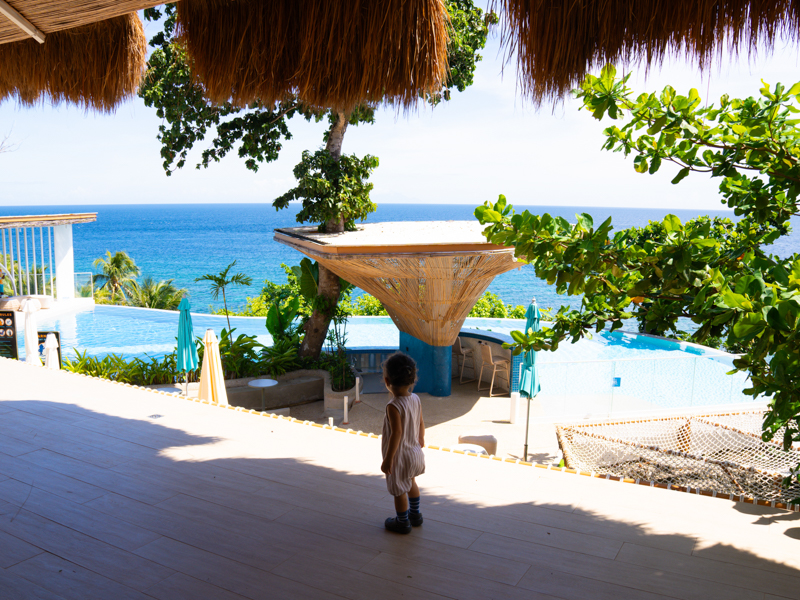 Child in light romper stands on a terrace overlooking a blue ocean and pool, with thatched shade structures and tropical plants nearby.