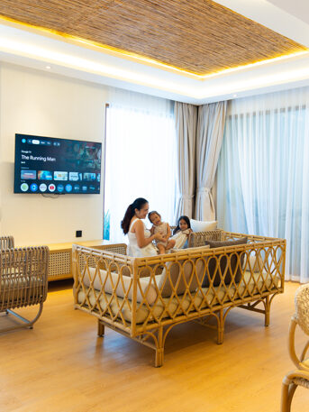Family of four sits on a woven rattan sofa in a bright living room with a wall-mounted TV and large windows on sheer curtains.