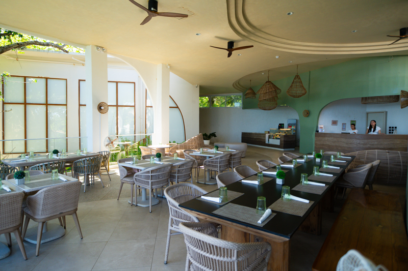 Sunlit restaurant interior with a long communal table, wicker chairs, and green glass cups set with napkins on each place.