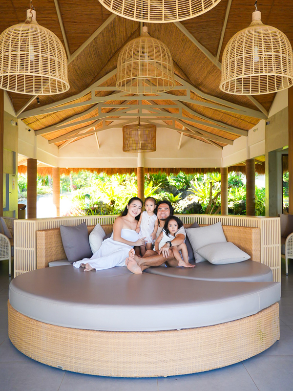 Family of four sitting on a round wicker sofa with gray cushions inside an open tropical pavilion with woven hanging lamps.