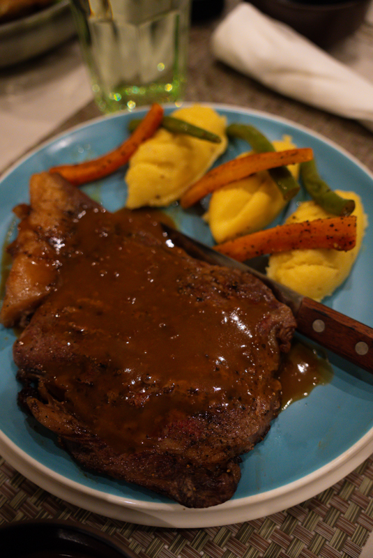 Plate with a large grilled steak covered in brown sauce, beside mashed potatoes and roasted carrots/green beans on a blue plate. A knife rests nearby.
