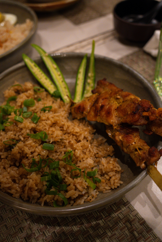 Plate of fried rice with chopped green onions, cucumber spears, and grilled chicken skewers on the side.