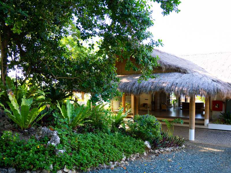 Tropical resort building with a thatched roof veranda surrounded by lush green plants and a gravel path.