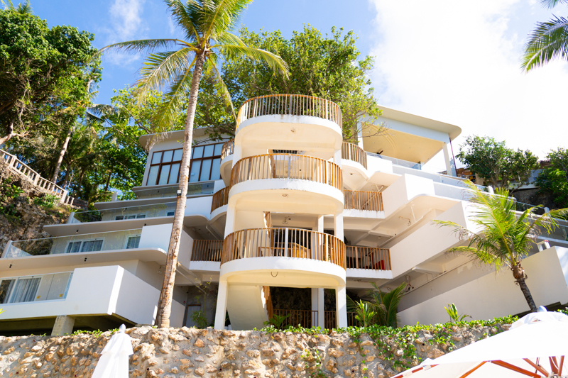White modern hotel with curved balconies surrounded by palm trees under a blue sky.