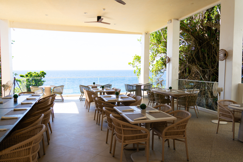 Open-air restaurant terrace with wicker chairs and tables set for dining, overlooking the ocean view.