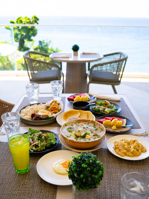 Outdoor seaside brunch table with a variety of dishes and fruit, overlooking the ocean and balcony seating in the background.