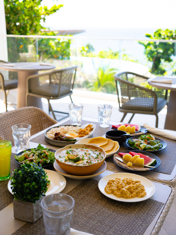 Outdoor patio table set for brunch with multiple dishes and a sea-view backdrop on a sunny day.