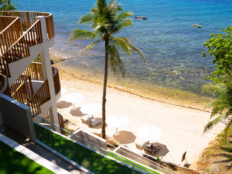Beach scene with white sand, palm trees, and white umbrellas by the shoreline; balconies on the left hint at a building exterior.