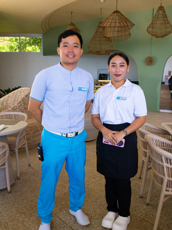 Two cafe staff stand side by side in a bright dining area with wicker chairs and hanging lamps in the background.