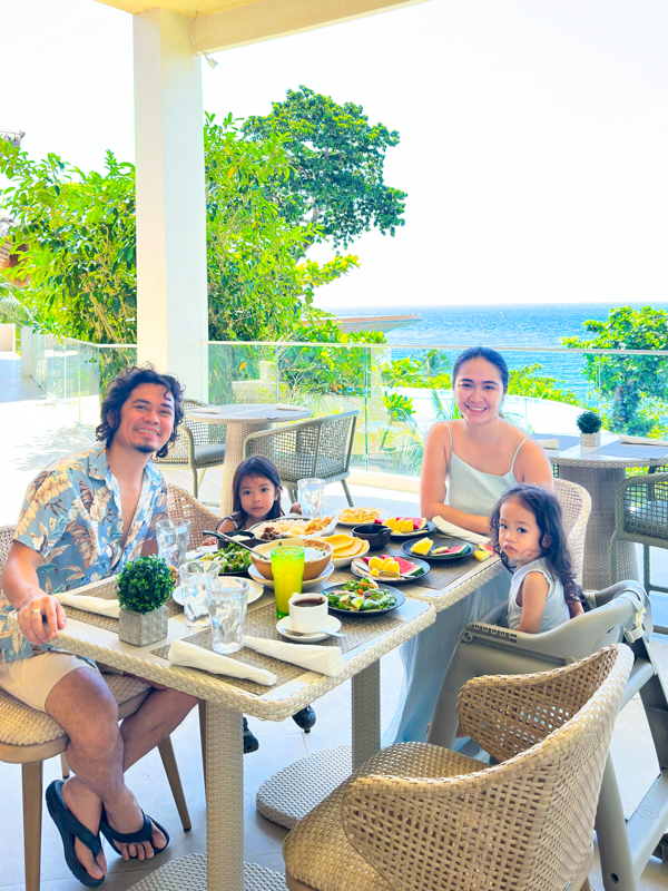 Family of four enjoying a sunny breakfast at an outdoor terrace with an ocean view.