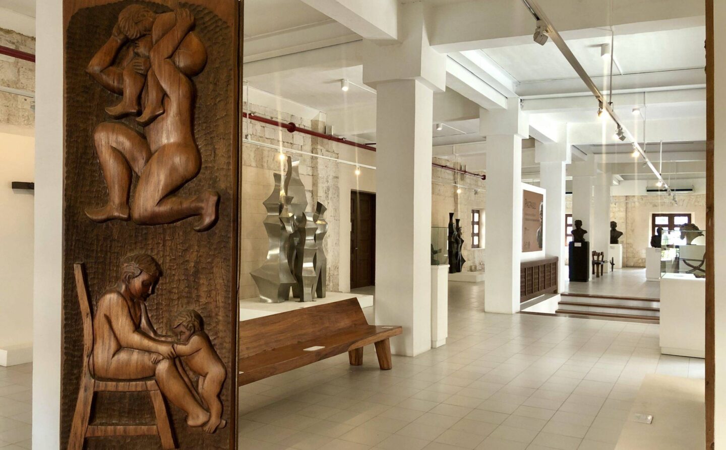 Museum interior with a large carved wooden panel and bench in the foreground, white pillars, and abstract metal sculptures in the background.