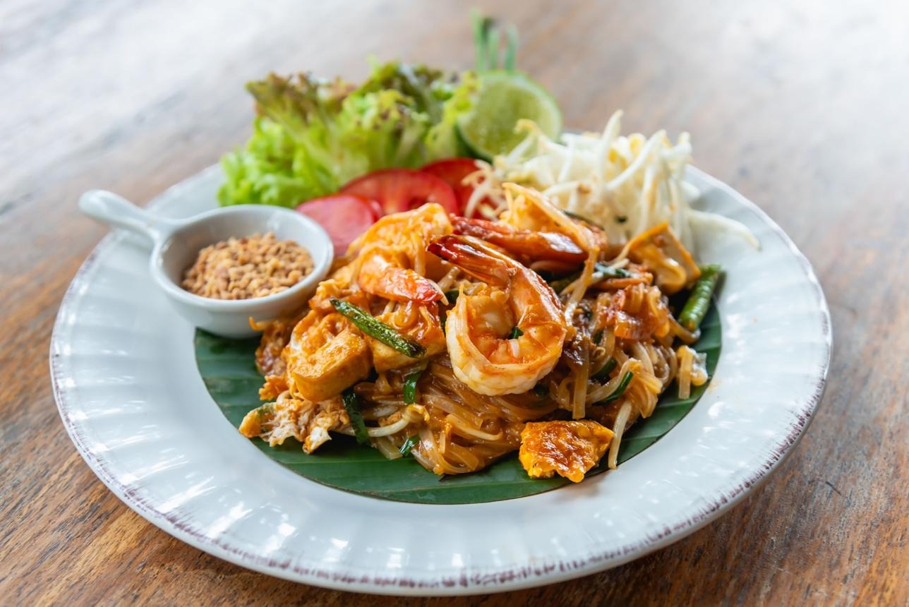 Pad Thai with shrimp and tofu on a white plate, with lime, peanuts, and a side salad on a banana leaf.