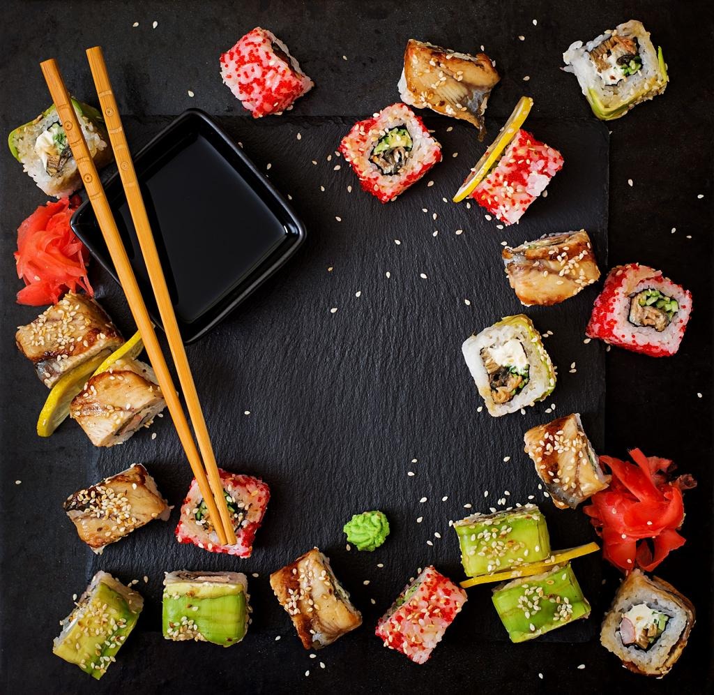 Assorted sushi rolls arranged in a circle on a dark slate, with a soy sauce dish, chopsticks, wasabi, and pickled ginger.