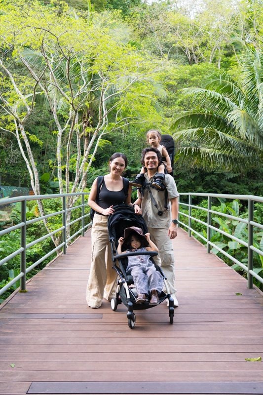 Family of four posing on a wooden bridge in a tropical forest; parents push a stroller with a toddler while a second child sits on dad's shoulders behind them.