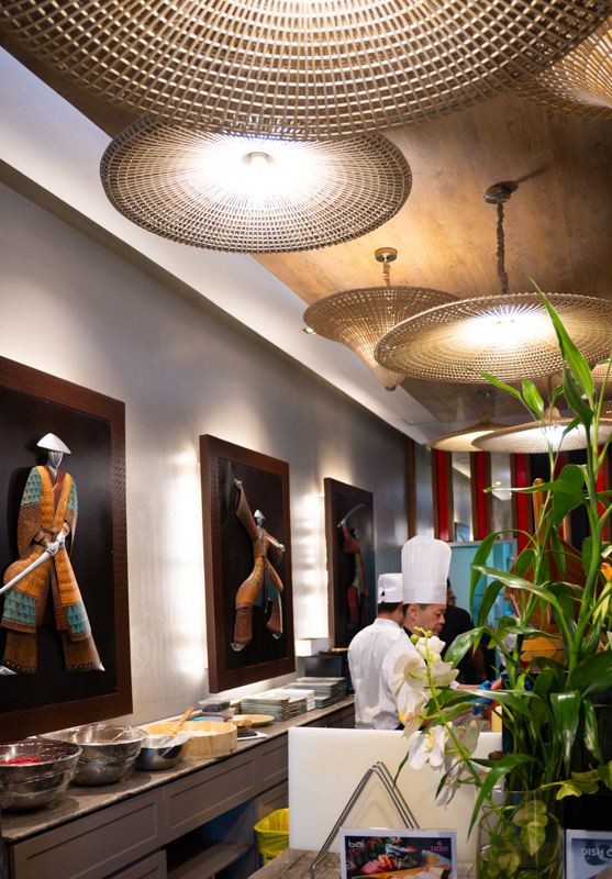 Restaurant kitchen area with chefs in white hats beside a long counter and framed clothing art on the wall, large woven pendant lights overhead.