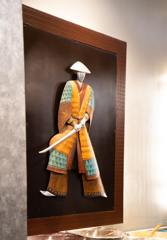 Colorful samurai figure in orange and teal armor, mounted on a dark display panel with a patterned brown frame, holding a curved sword.