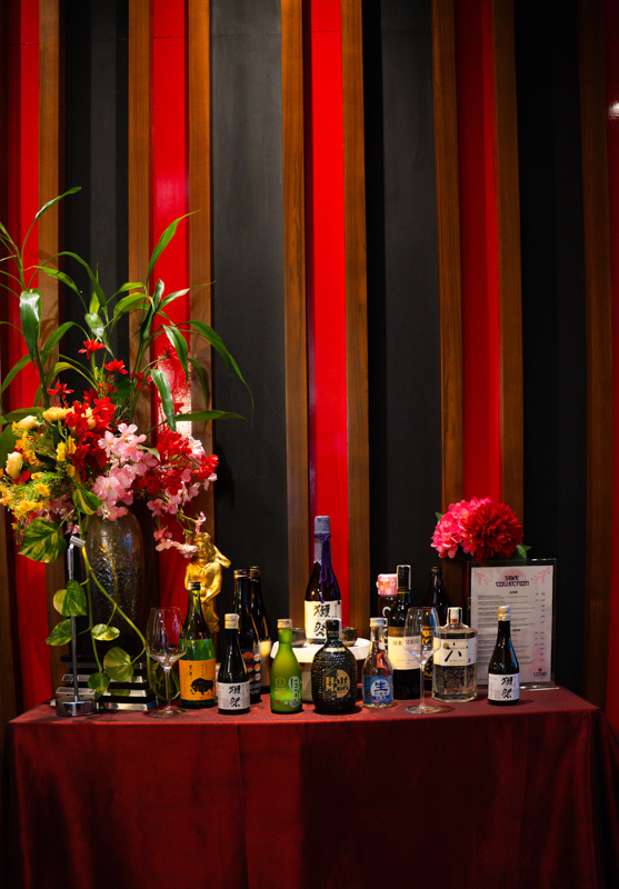 Table with assorted bottles and flowers against a striped colorful wall, likely a beverage display for a celebration.