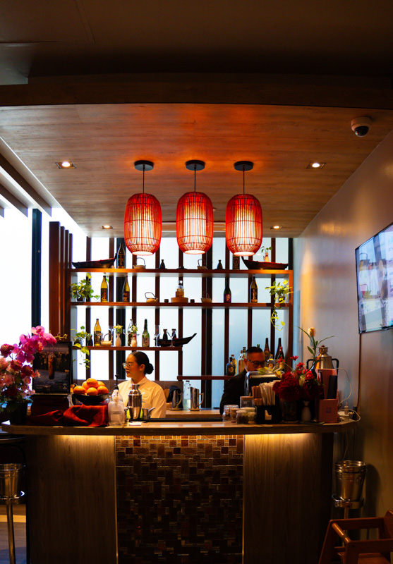 Interior bar scene with three red pendant lamps above a curved wooden counter and bottles on shelving behind two staff members.