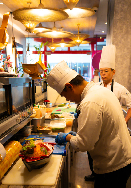Two chefs in white uniforms and tall hats work at a sushi bar; one slices sashimi onto a bowl at the counter while the other watches nearby.