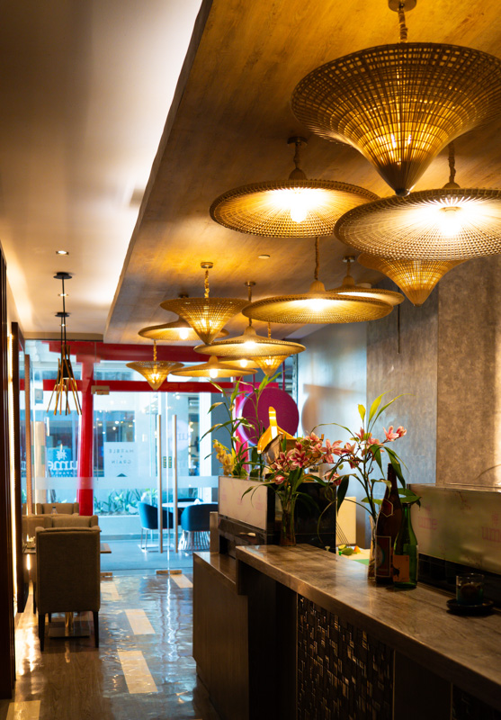 Interior of a stylish lounge with warm pendant lights over a dark wooden bar and floral arrangements along the counter, leading to glass doors at the back.