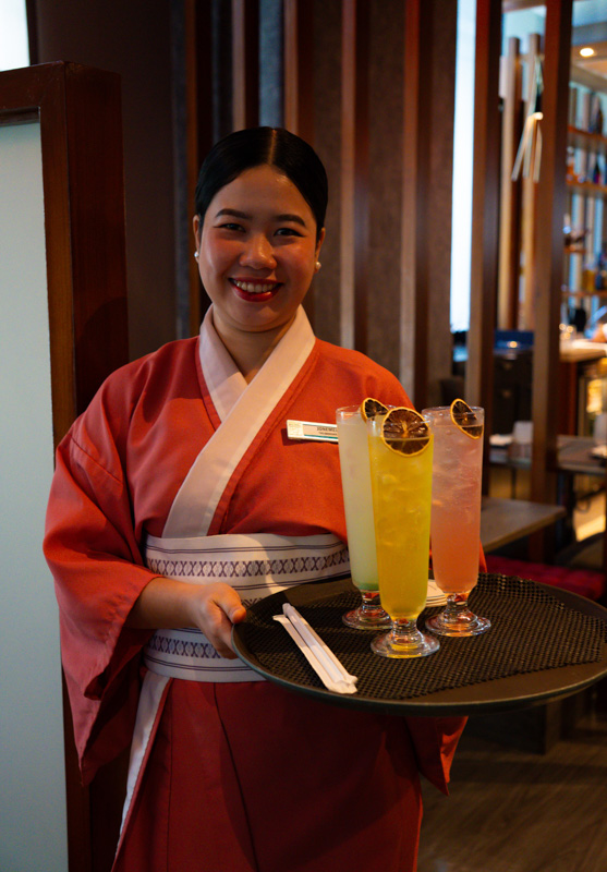 Smiling server in a red kimono-style uniform presenting a tray with three colorful cocktails garnished with citrus slices in a restaurant setting.