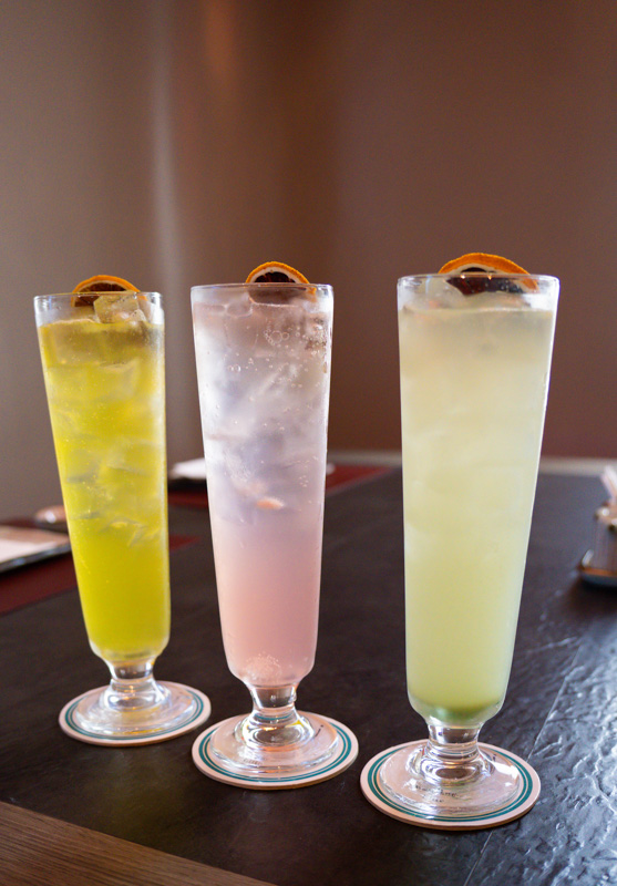 Three tall cocktails with orange garnish on a dark bar; left is yellow-green, middle pink, right pale green.