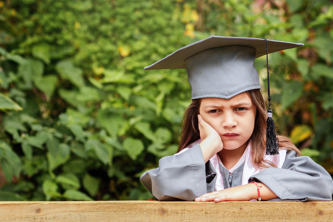 Young girl in a gray graduation cap and gown rests her head on her hand outdoors, looking bored.