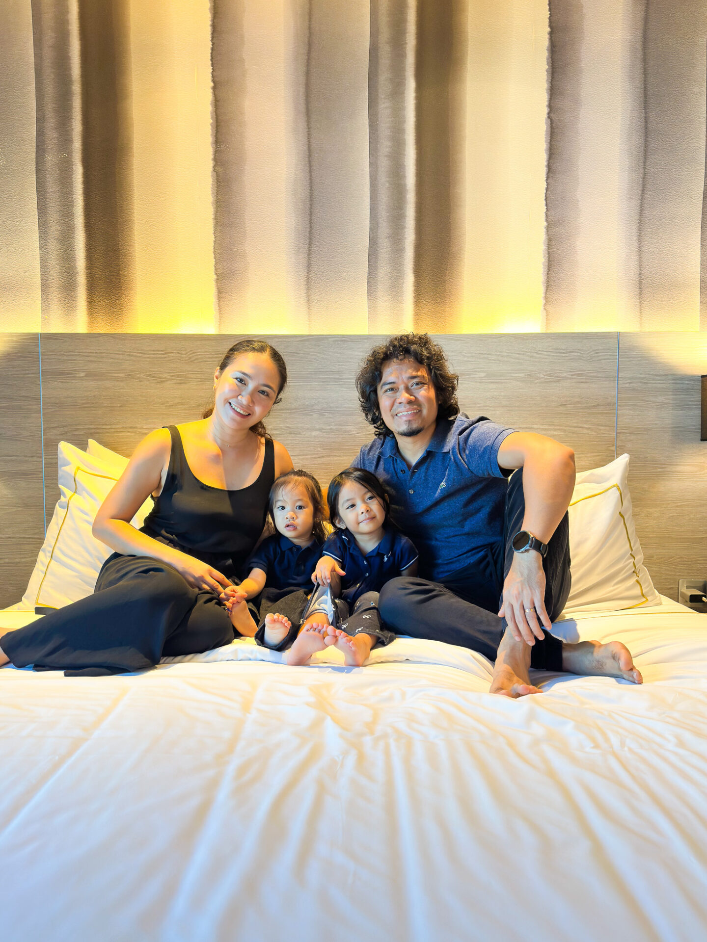 Family of four sits together on a hotel bed, smiling at the camera with two young children between them.