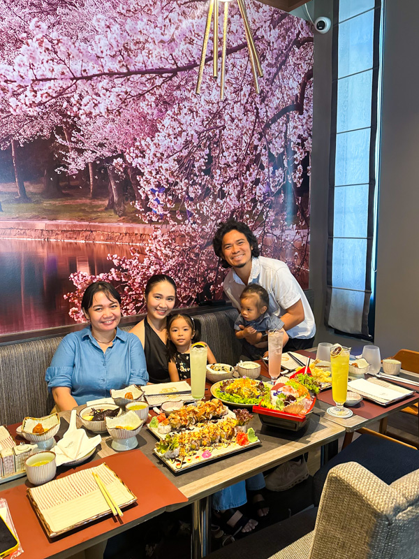 Family of five at a restaurant table, smiling; cherry-blossom mural behind them and assorted dishes on the table.