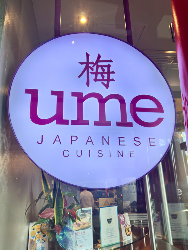 Circular illuminated sign with the word 'ume' and Japanese character 梅 above, 'JAPANESE CUISINE' below, displayed in a restaurant window.