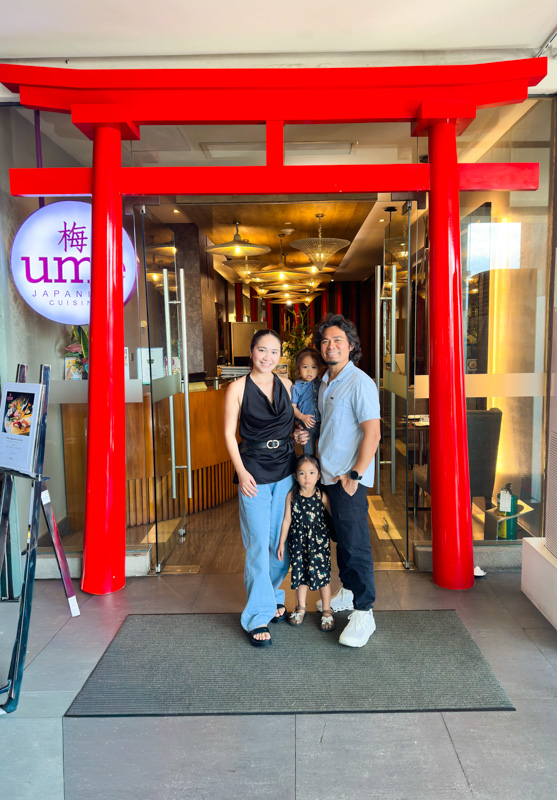 Family posing under a red torii gate at the entrance of a Japanese restaurant, with a mother, father, and two children in view as they smile for the photo.