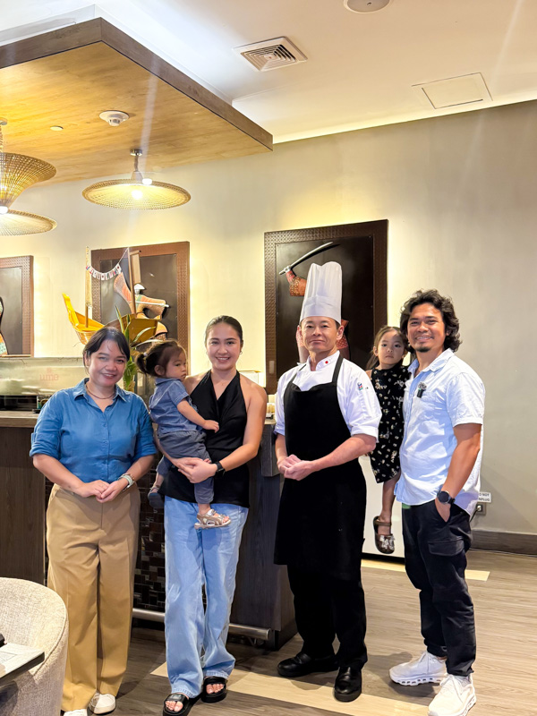 Six people in a restaurant posing for a photo: a chef in a tall white hat and black apron with a family.