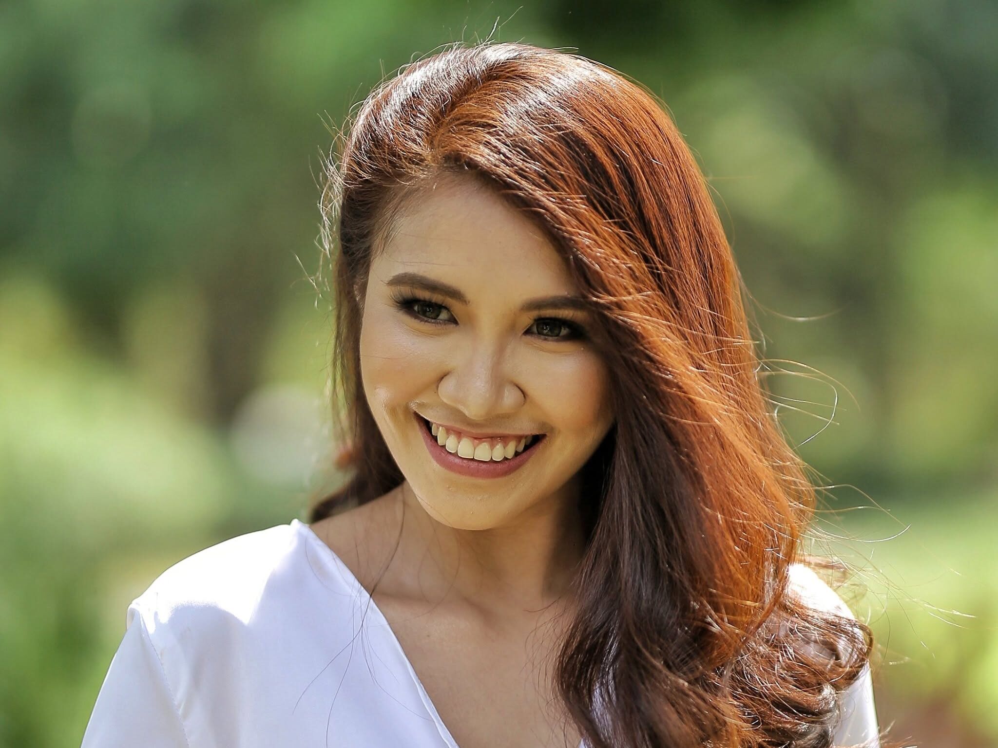 Close-up of a smiling woman with long brown hair wearing a white blouse outdoors.