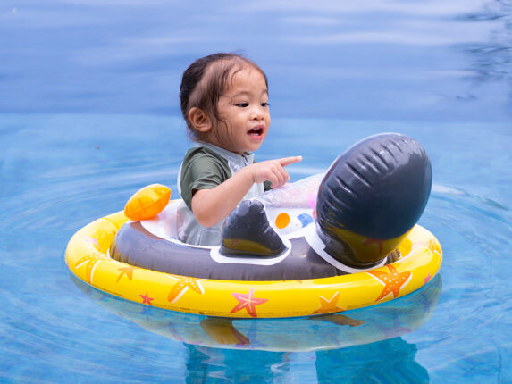 Toddler in a yellow star-pattern float in a swimming pool, pointing at something off‑camera.