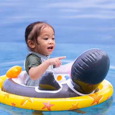Toddler in a yellow star-pattern float in a swimming pool, pointing at something off‑camera.