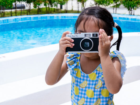 Young girl in a lemon-printed swimsuit holds a vintage camera up to her face by a pool, as if taking a photo of the scene behind her.