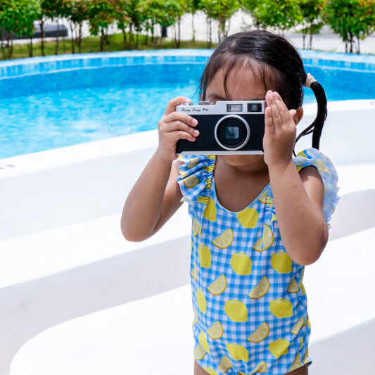 Young girl in a lemon-print swimsuit holds a camera up to her face by a bright blue pool area.